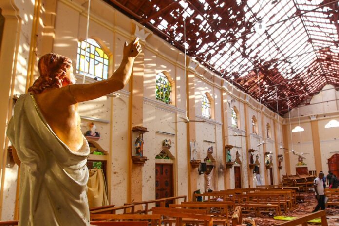 Statue of Jesus with raised arm inside a damaged church, sunlit broken roof and debris on pews.